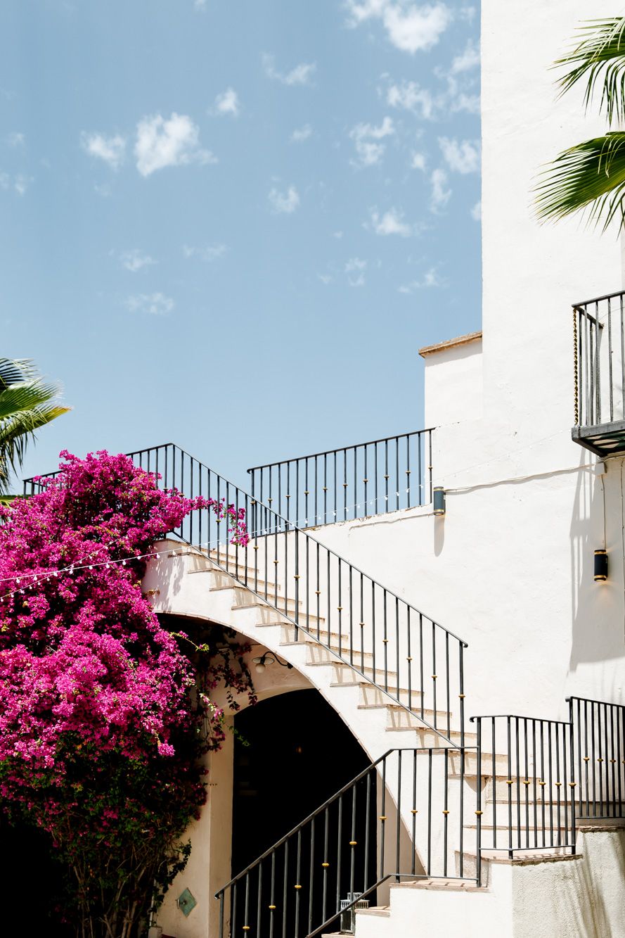 Masia Victoria wedding venue with curved staircase, vibrant pink bougainvillea, black iron railings, and palm trees under blue sky.