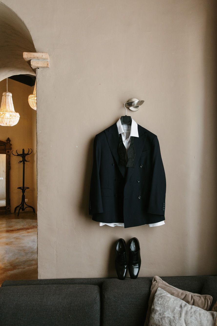 Dark suit jacket and white dress shirt hanging on wall hook above black Oxford shoes on a sofa, wedding attire display.