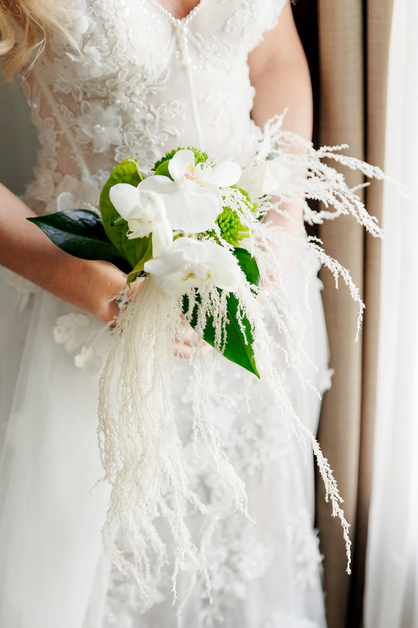 A delicate white bridal bouquet with green leaves and wispy accents is held against a lace wedding dress.