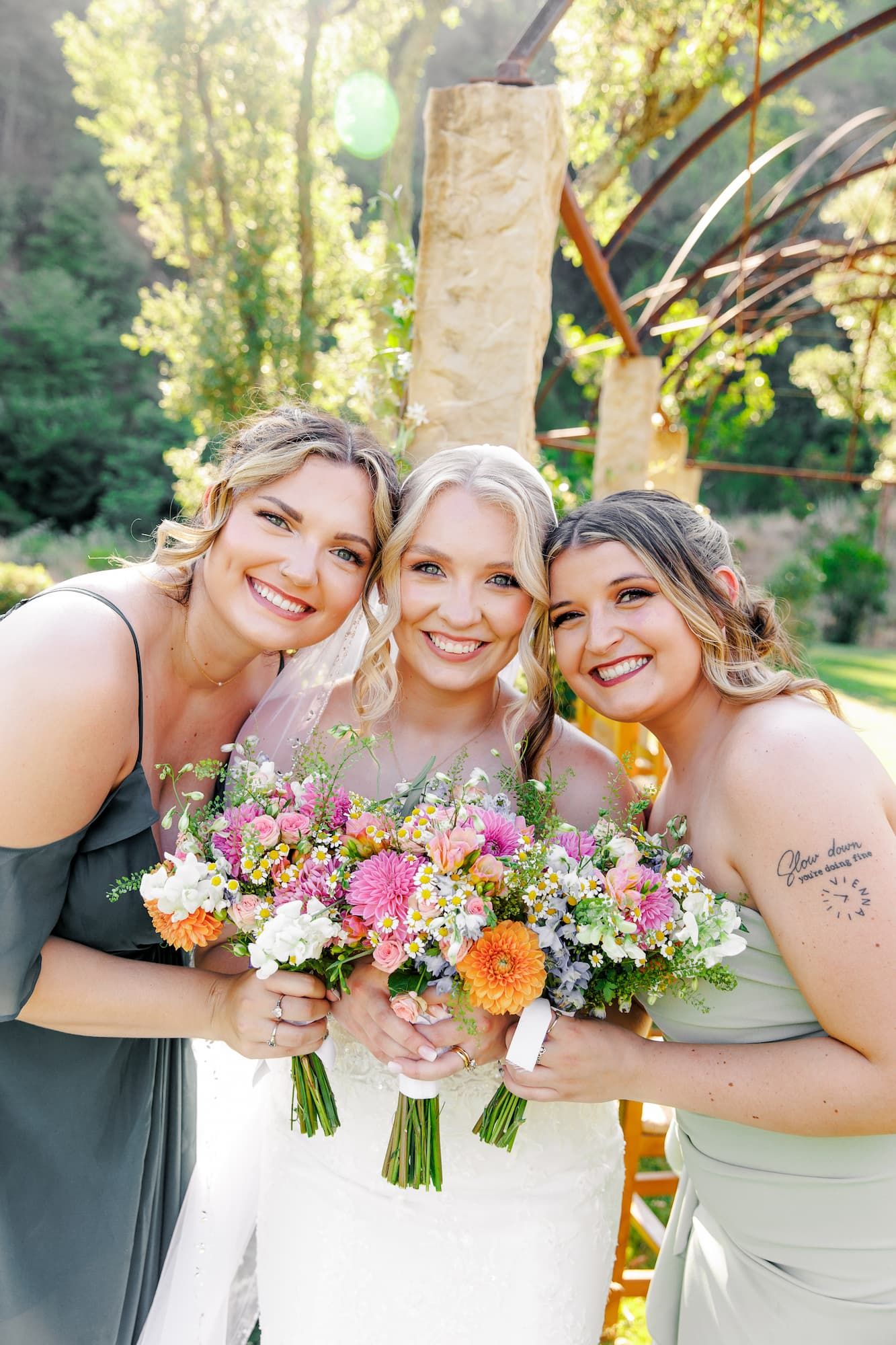 Three bridesmaids pose together with colorful bouquets of pink and orange flowers at an outdoor wedding.