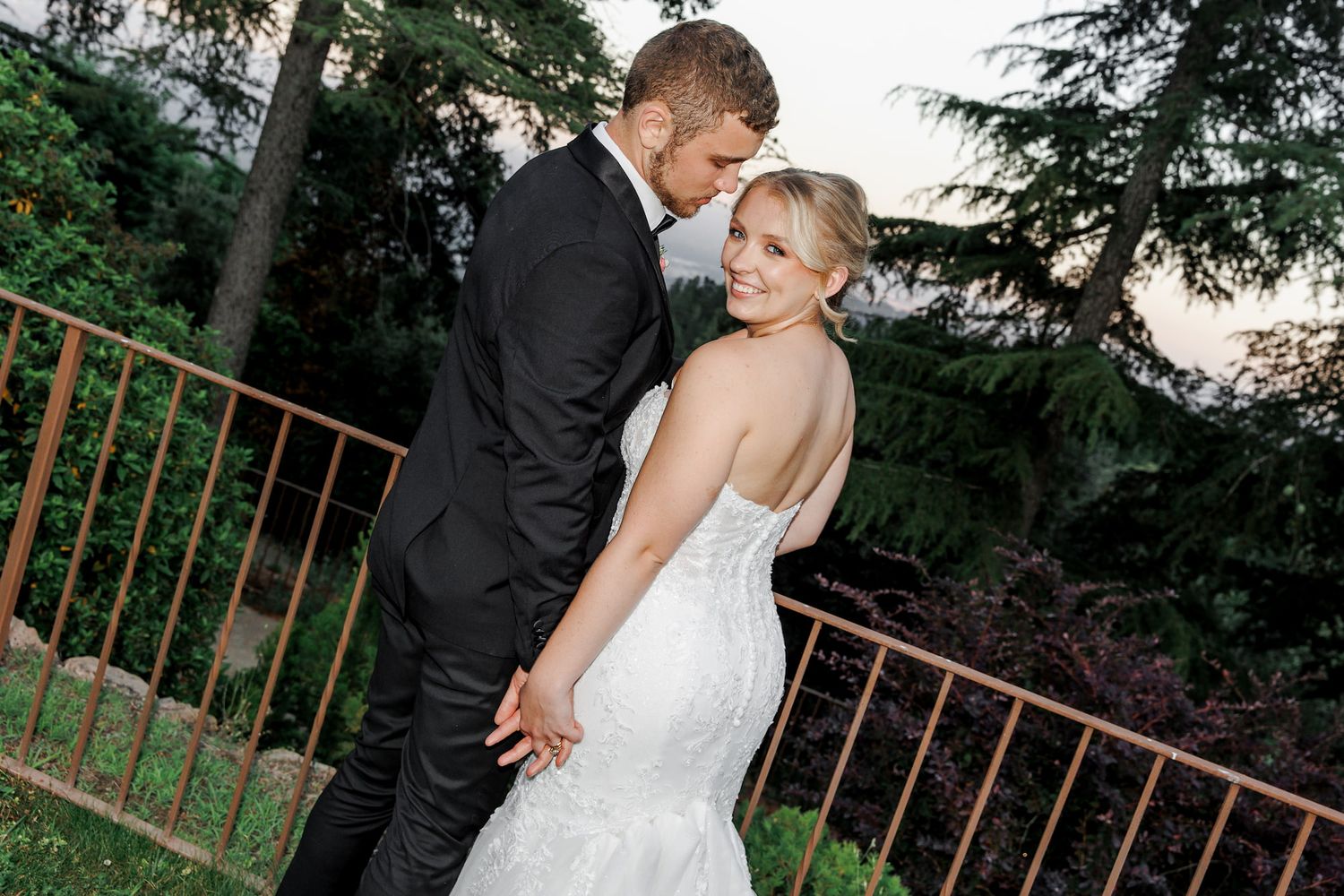 A romantic moment between newlyweds posing against an outdoor railing with trees in the background.