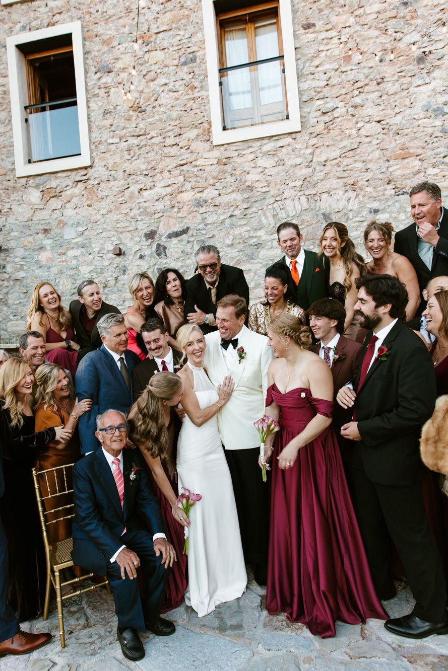 Smiling wedding guests in formal attire gather around a bride in a white gown and groom in a white jacket outside a rustic stone building.