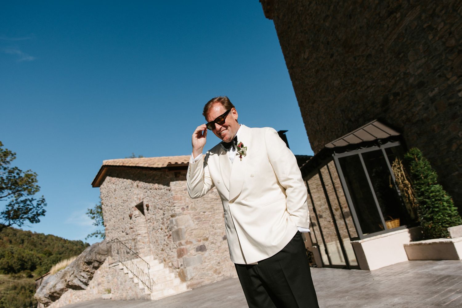 Man in a white dinner jacket and bow tie adjusts his sunglasses on a sunlit stone terrace beside a rustic building under a clear blue sky.
