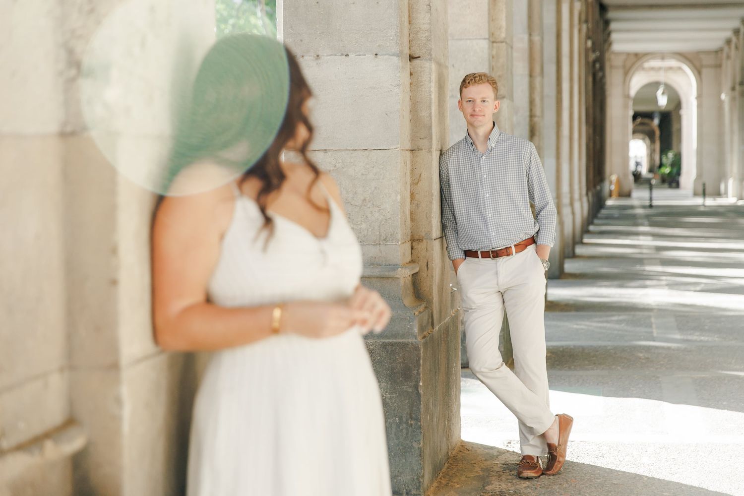 Couple poses in elegant stone colonnade, woman in white dress foreground, man in gray shirt leaning against pillar.
