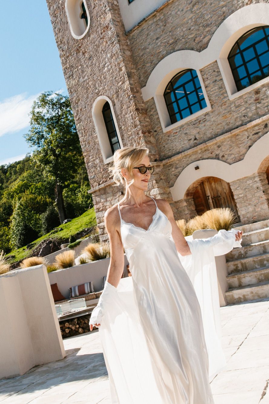Woman in sunglasses and a flowing white satin gown with gloves poses on a terrace beside a rustic stone tower with arched windows.