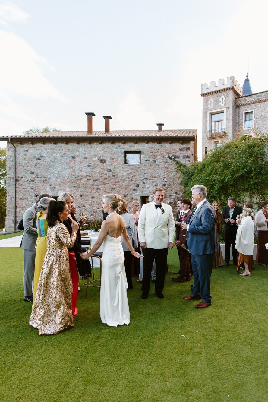 Wedding guests in formal attire mingle on a manicured lawn outside a rustic stone villa with a castle-style tower.