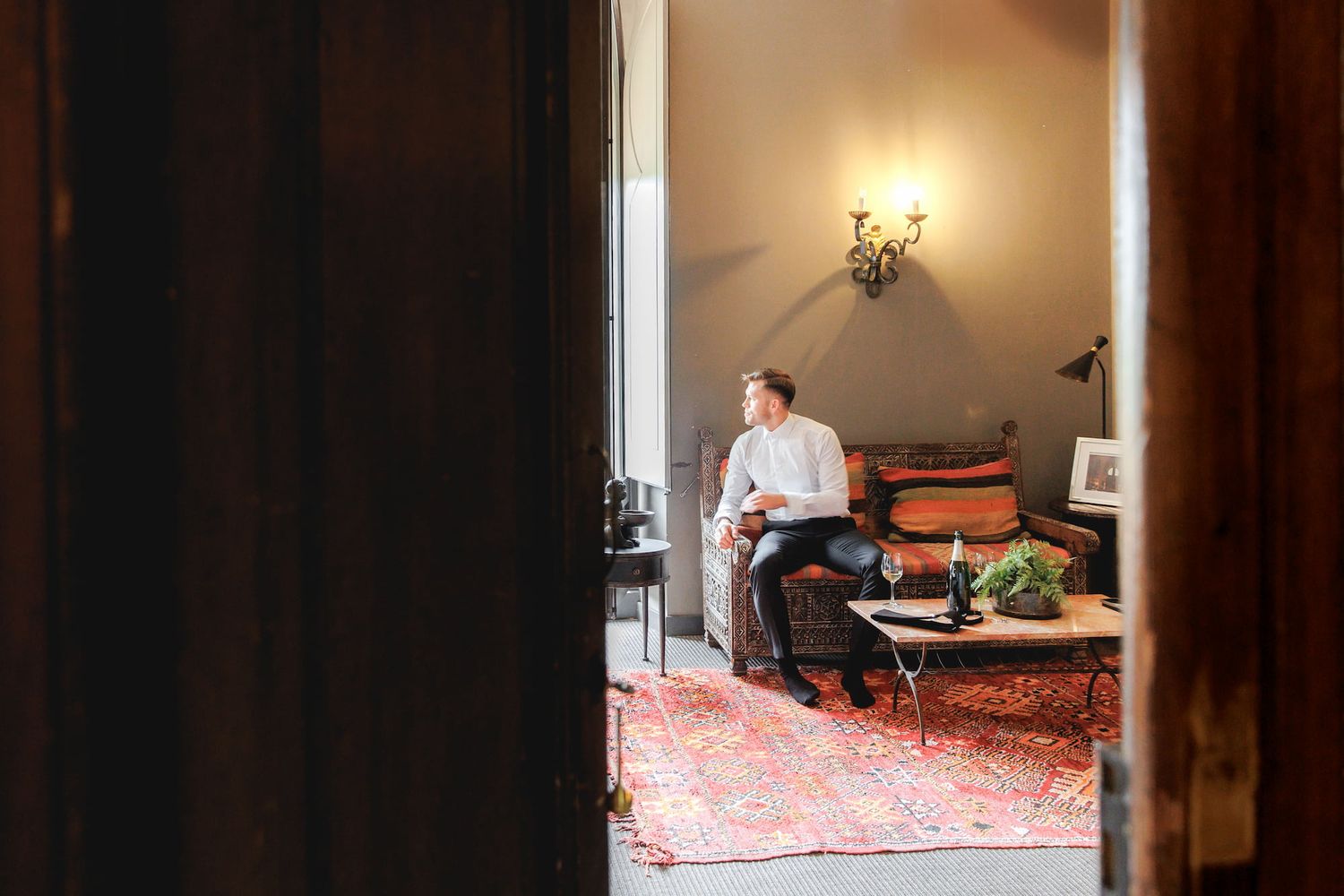 Person sitting on a sofa in a warmly lit living room with a red rug, viewed through an open doorway.