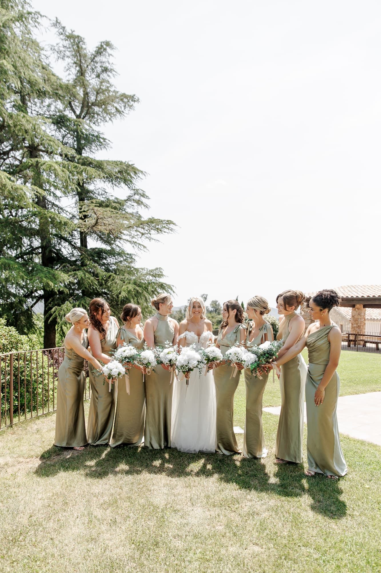 Bride in white gown poses with bridesmaids in sage green dresses holding white bouquets during the wedding in Spain