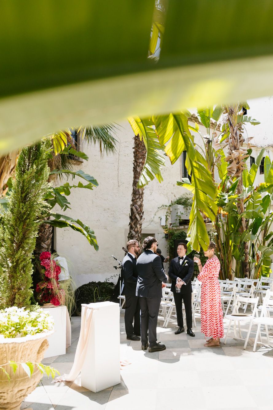 Outdoor wedding venue with tropical plants, white chairs, and guests in formal attire conversing near a white stucco wall.
