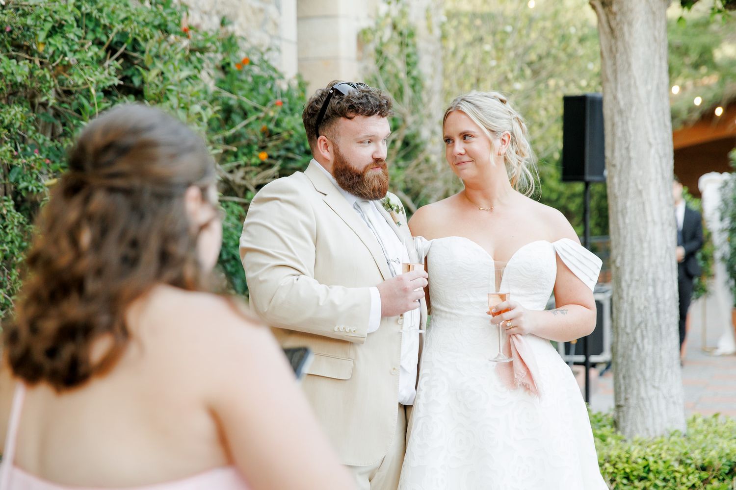 Bride in strapless white gown and groom in tan suit share a moment outdoors at elegant wedding reception.