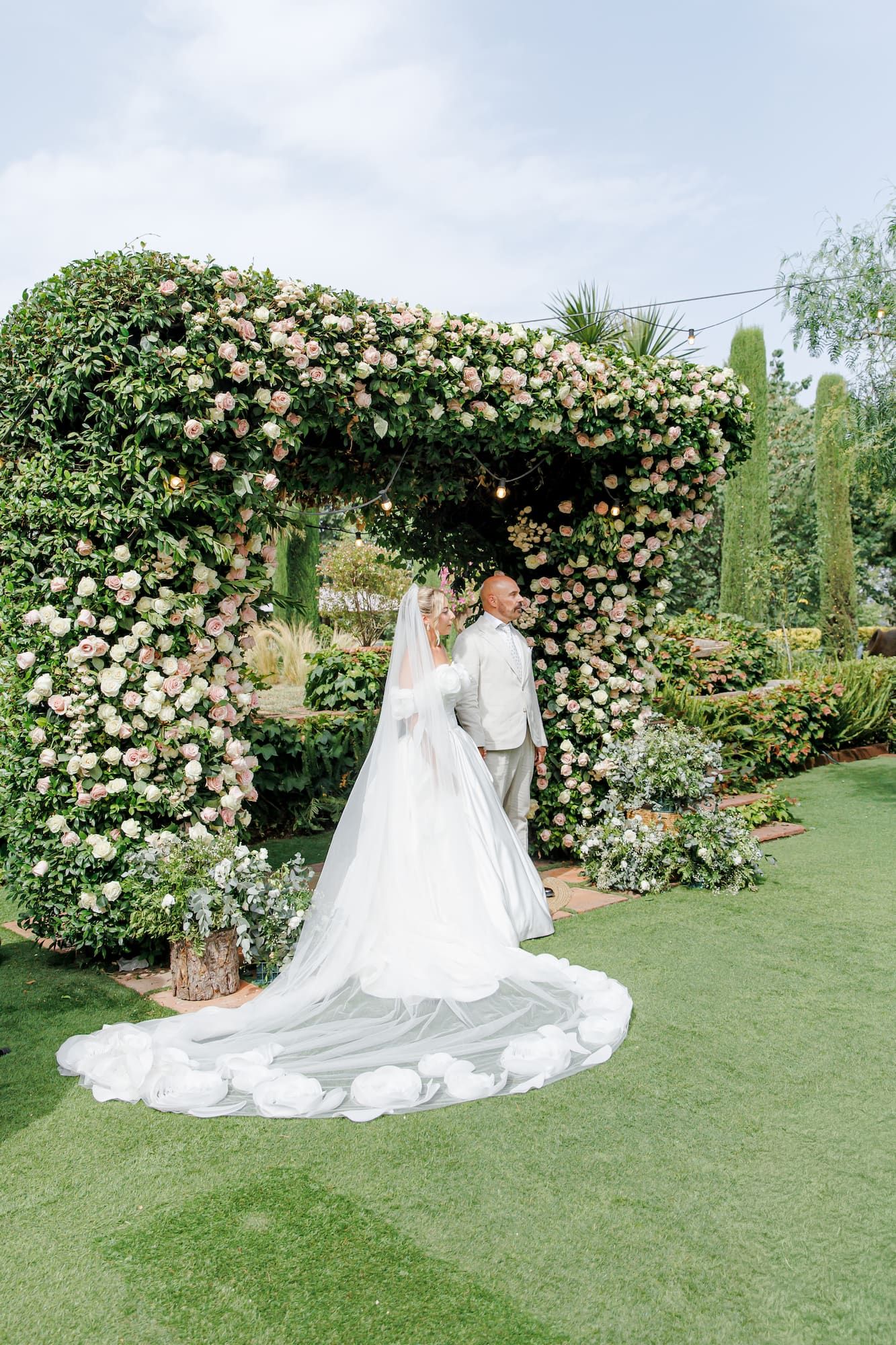 Bride and groom stand beneath a lush floral arch with pink blooms in a garden wedding ceremony.
