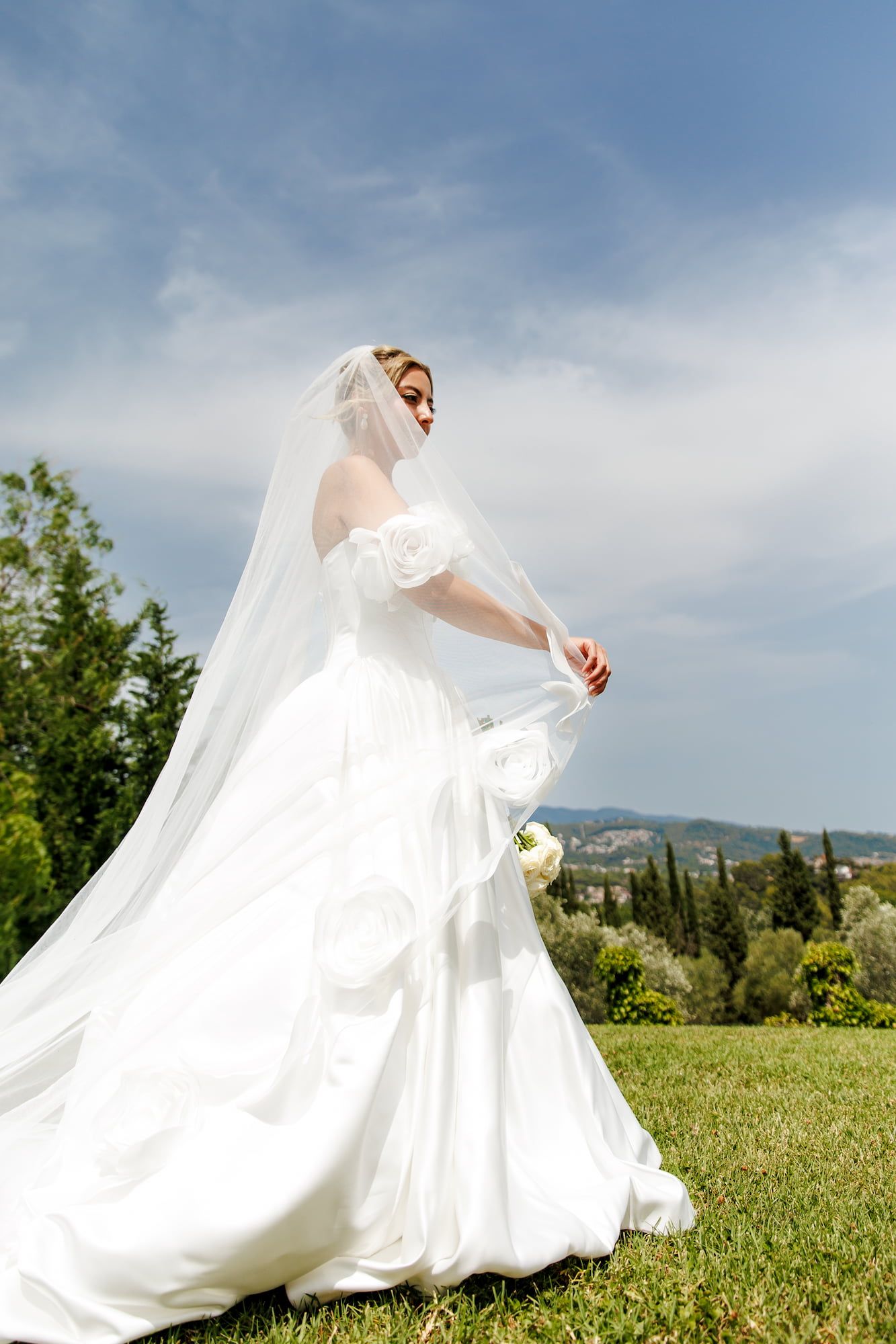 Elegant bride in white gown and veil stands on green lawn with scenic landscape and blue sky backdrop.