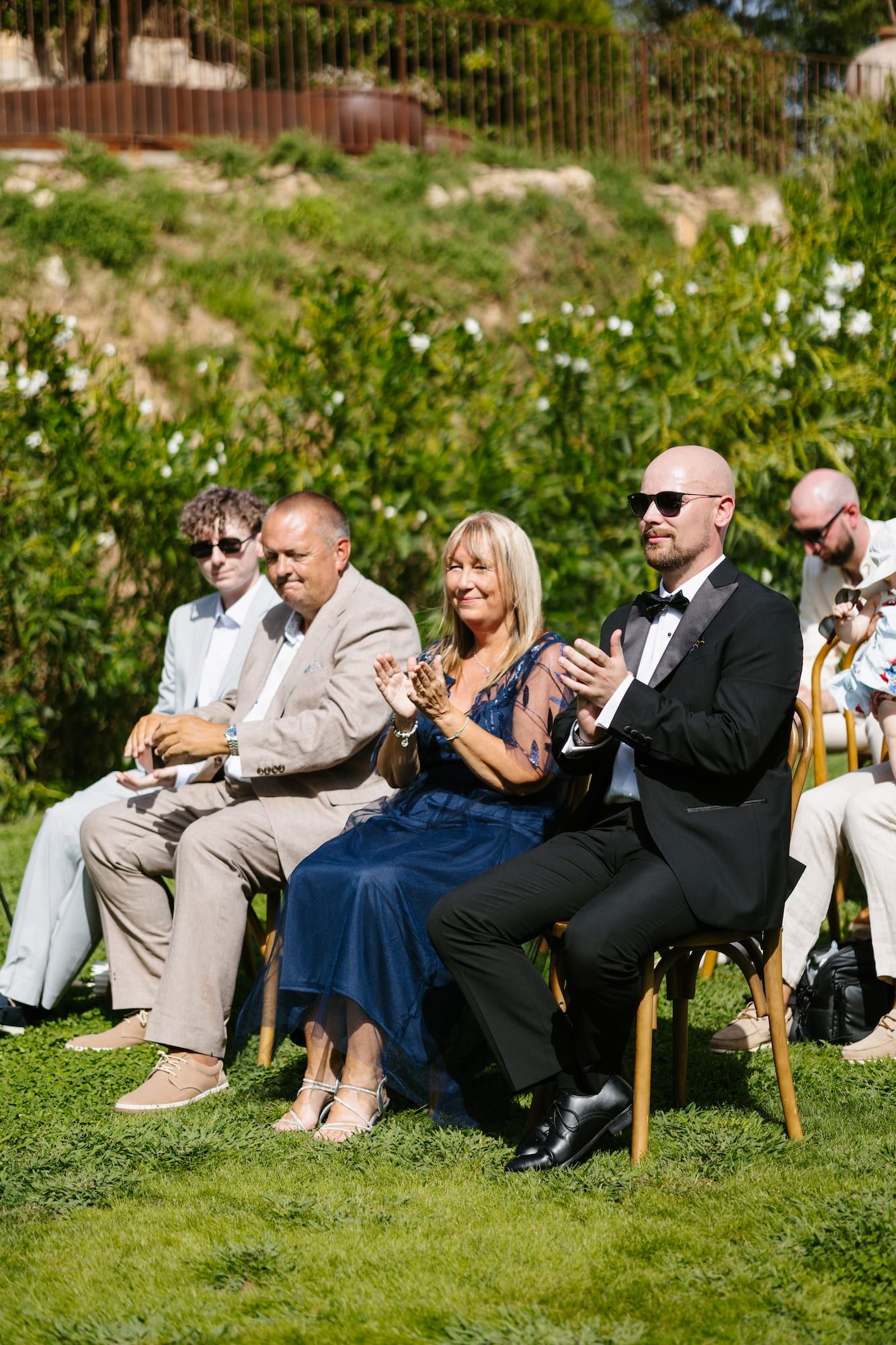 Wedding guests in formal attire clap and smile while seated outdoors during a sunny garden ceremony.