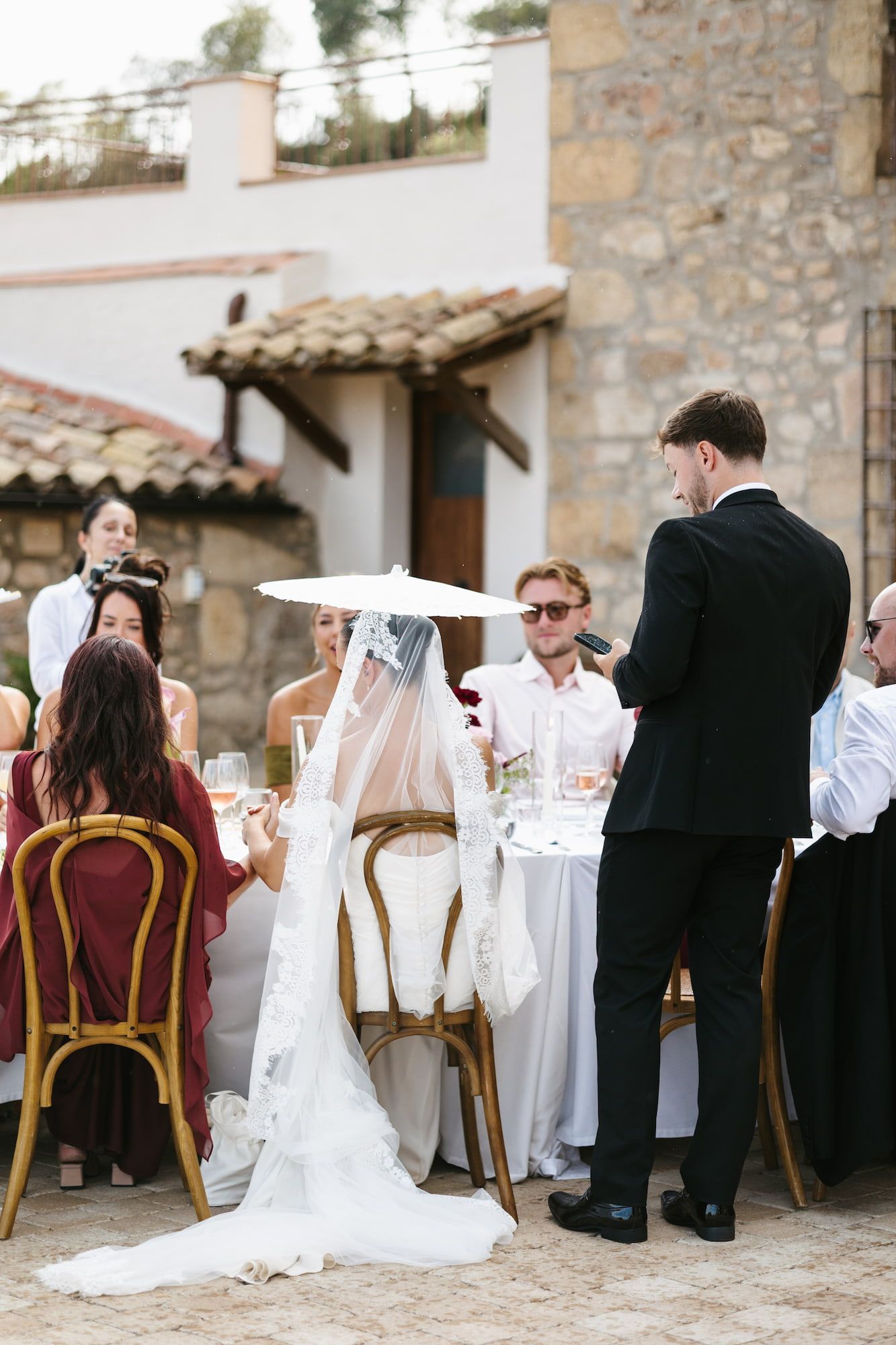 Outdoor wedding reception at a rustic stone villa, bride in white veil seated as groom gives a heartfelt speech.