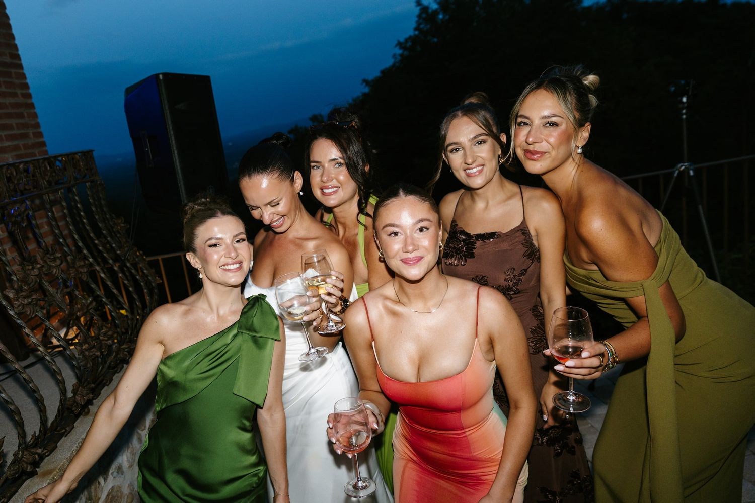 Six women in elegant dresses smile and pose together at an outdoor evening celebration.