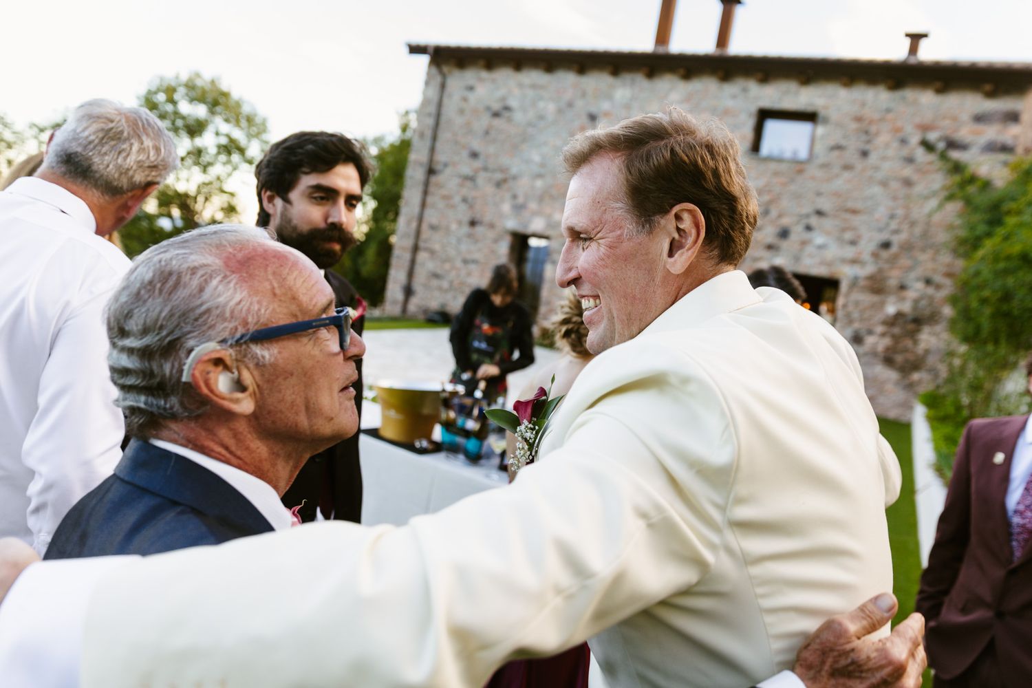 Two men in suits embrace warmly at an outdoor wedding celebration with guests and a rustic stone building in the background.