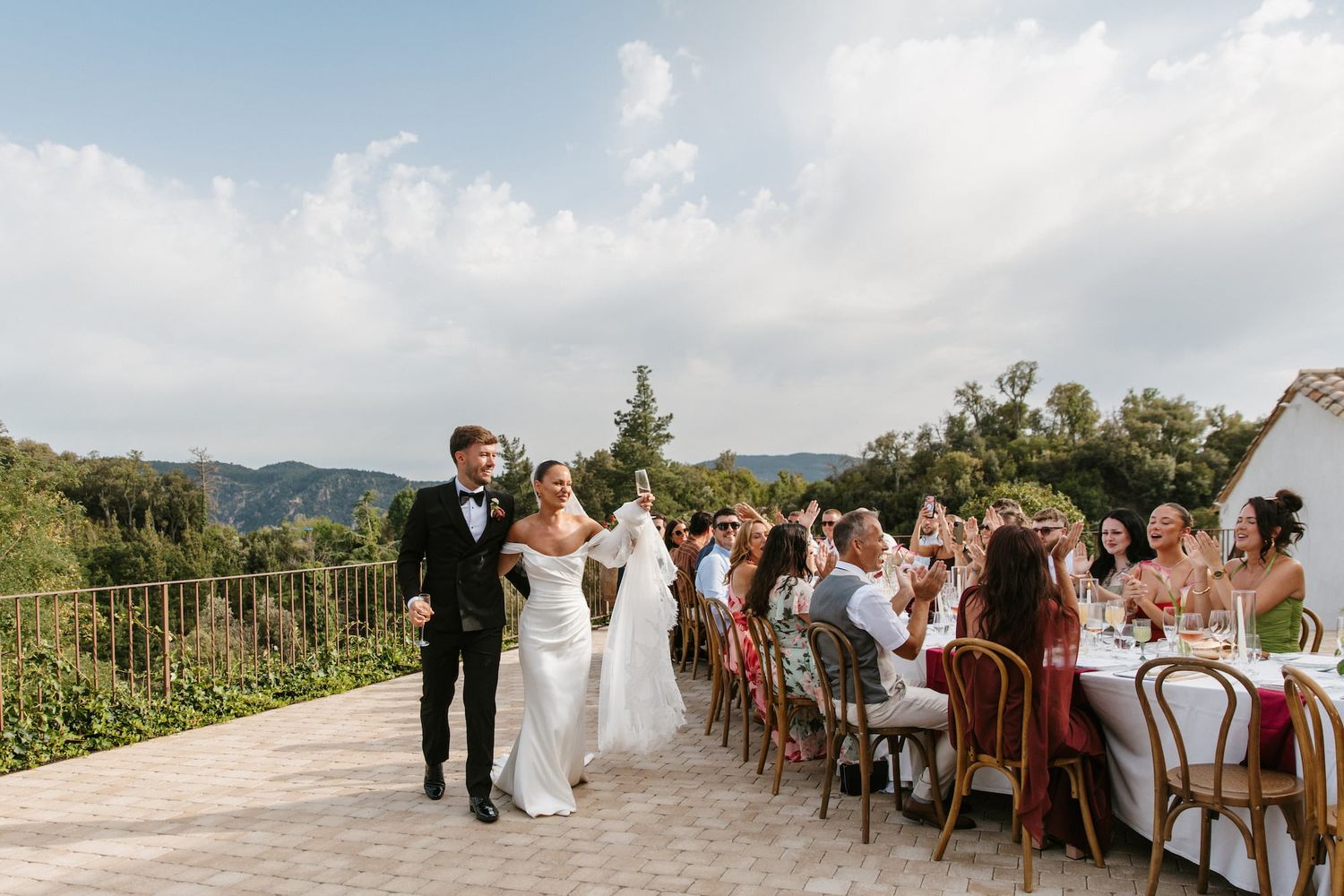 Bride and groom walk between reception tables outdoors with mountain views at a sunny Italian wedding celebration.