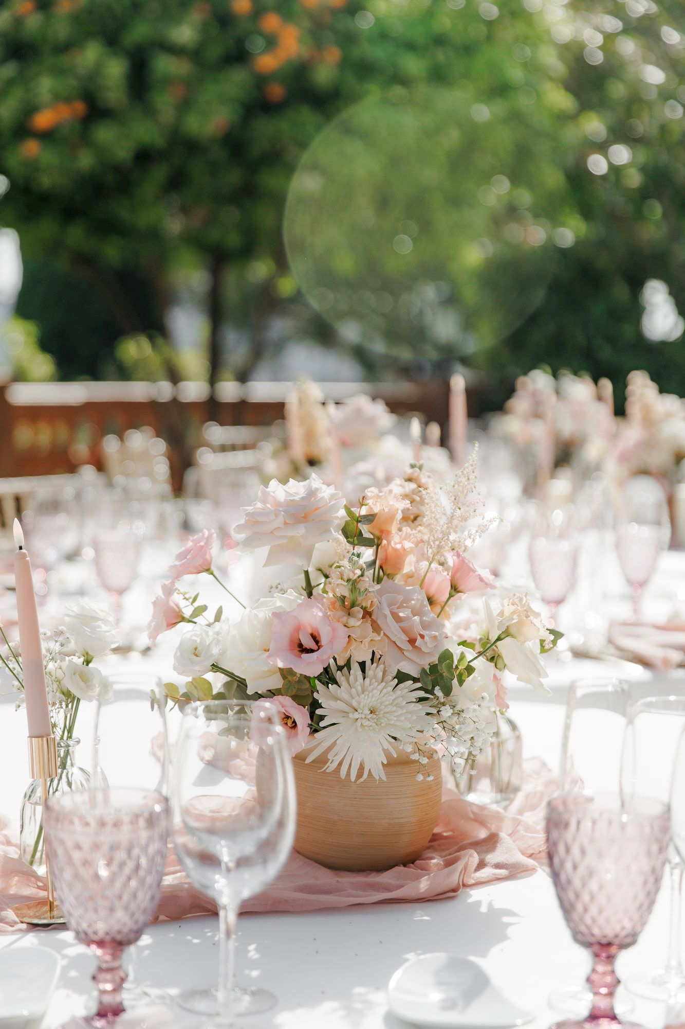 Elegant outdoor wedding table with pink floral centerpiece, blush glassware, and white linens under lush green trees.