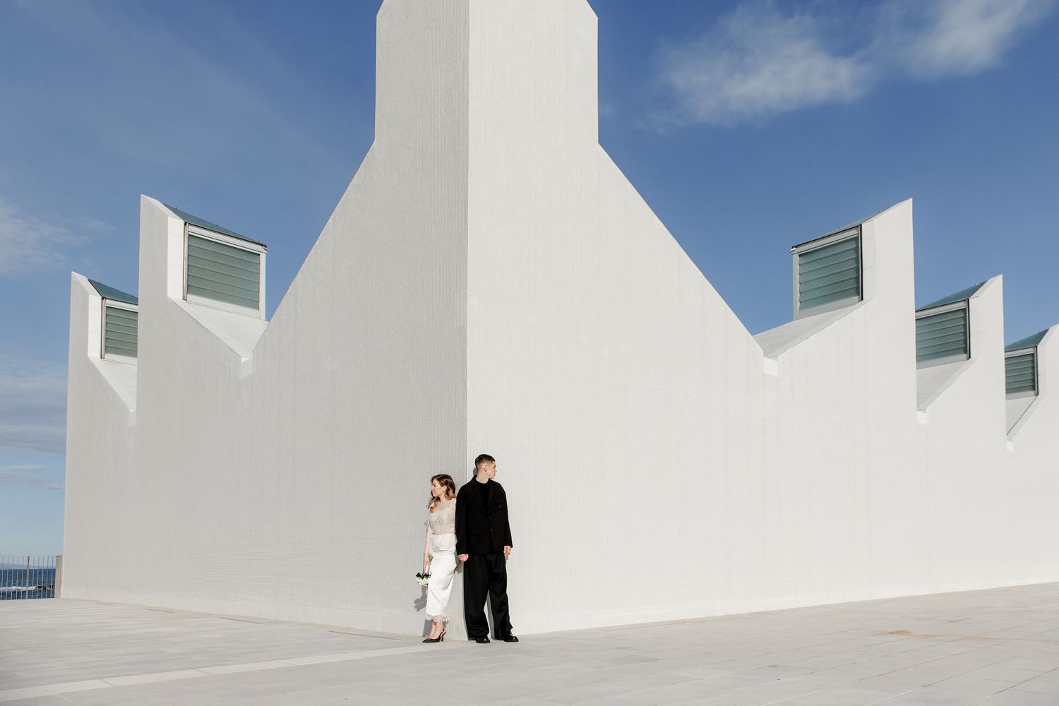 Couple in formal attire standing against a white architectural wall under a blue sky.