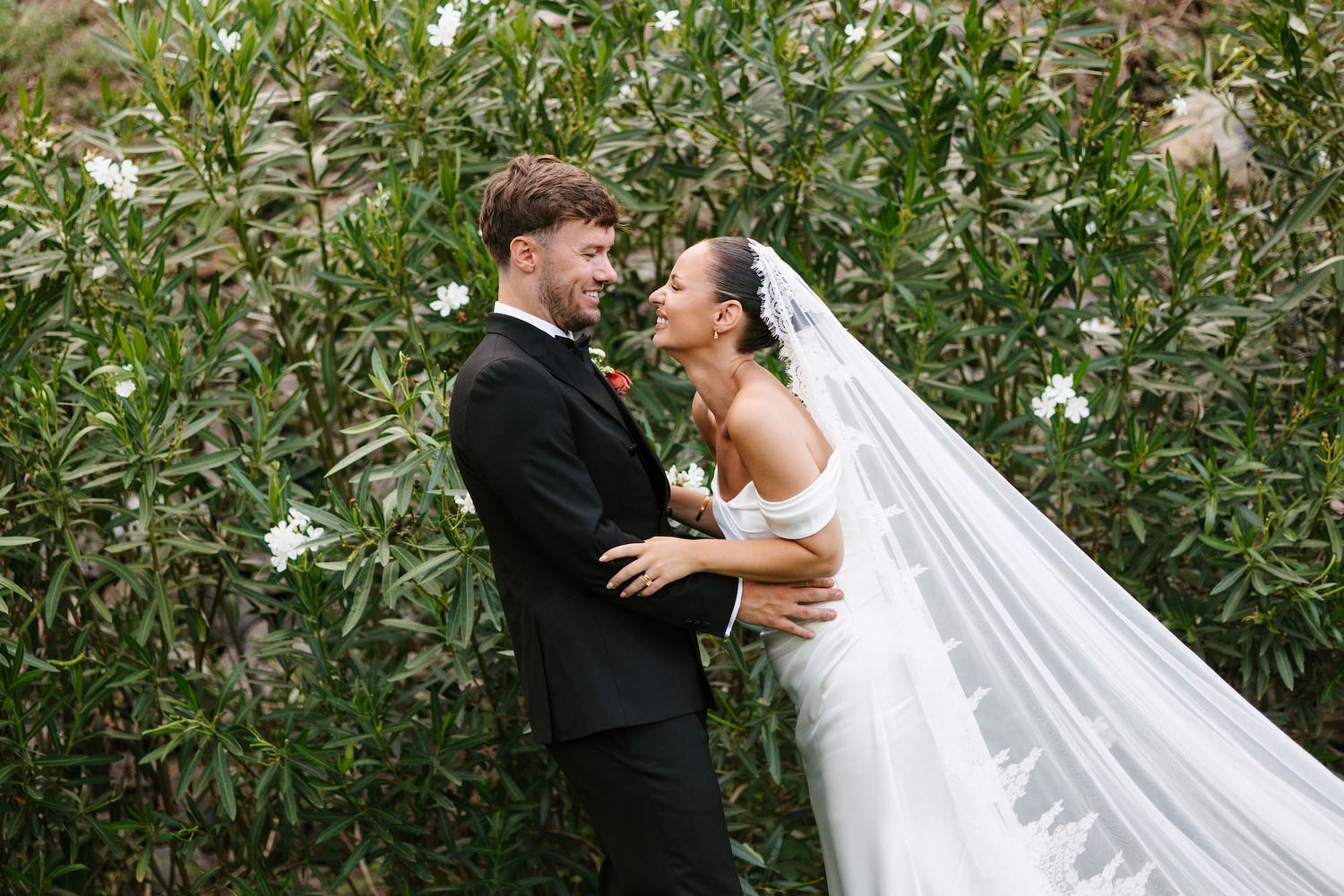 Bride and groom share a romantic moment surrounded by lush green foliage and white flowers on their wedding day.