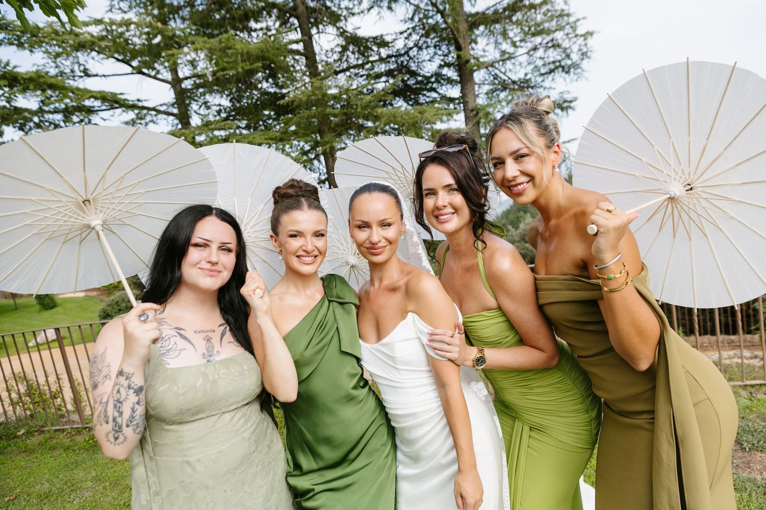 Bride in white dress with four bridesmaids in green dresses holding white parasols outdoors at a wedding.