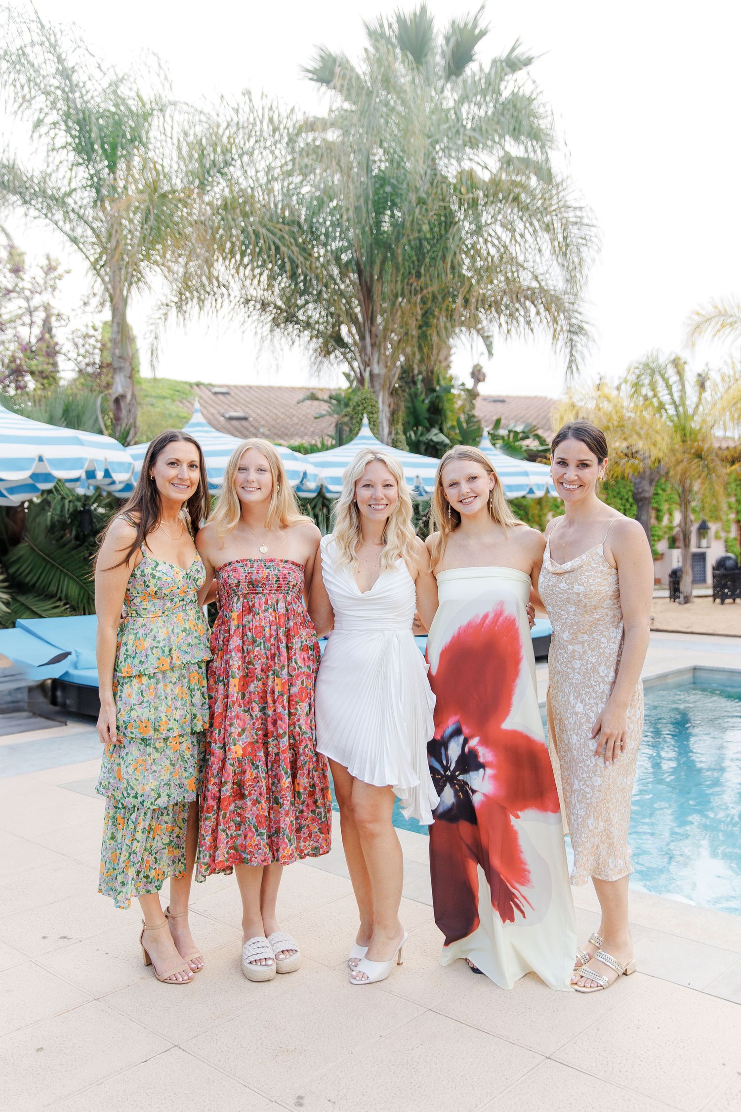 Five women in colorful summer dresses posing together poolside with palm trees during a rehearsal dinner at Gran Villa Rosa