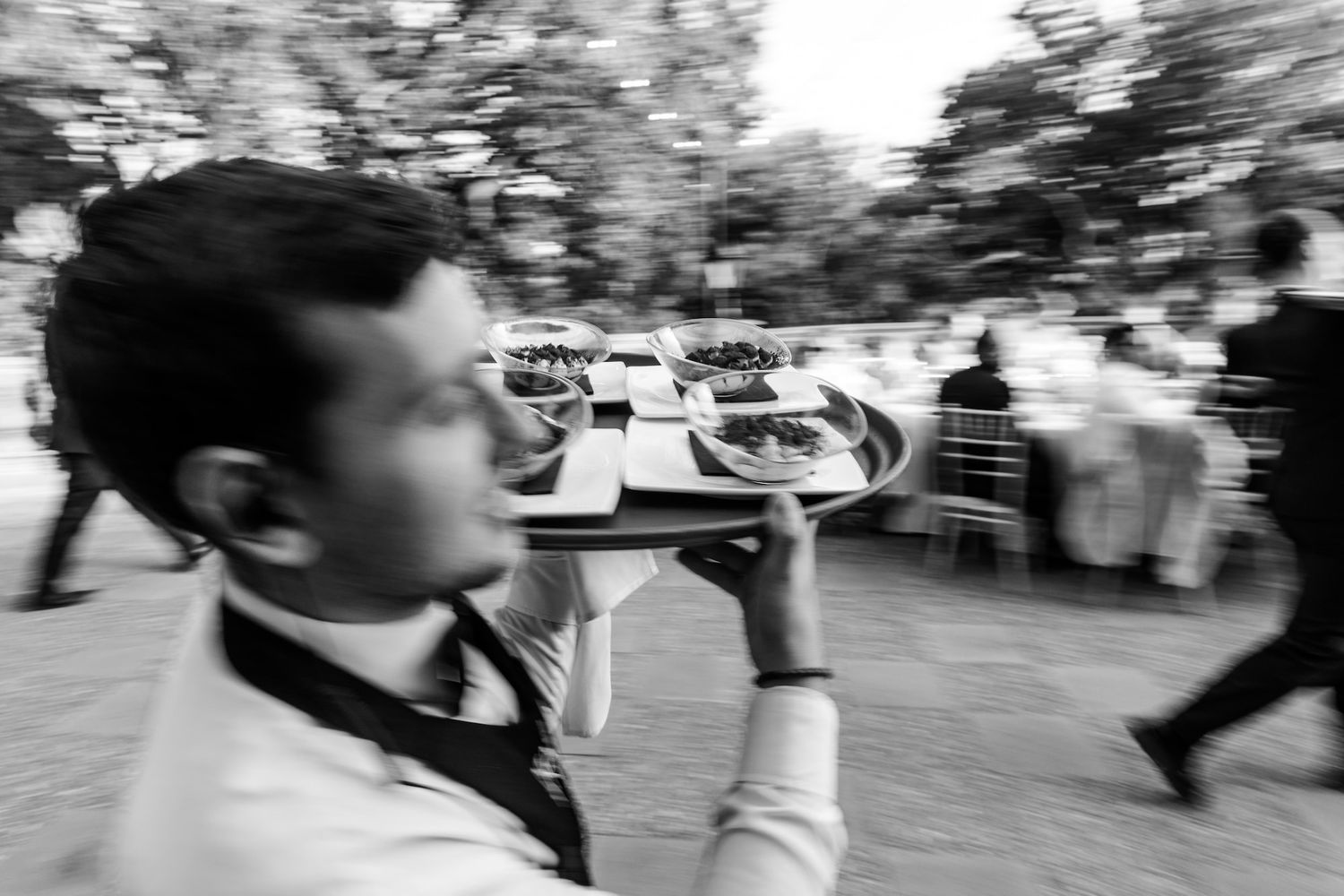Black and white photo of a waiter carrying a tray of food at an outdoor catering event.