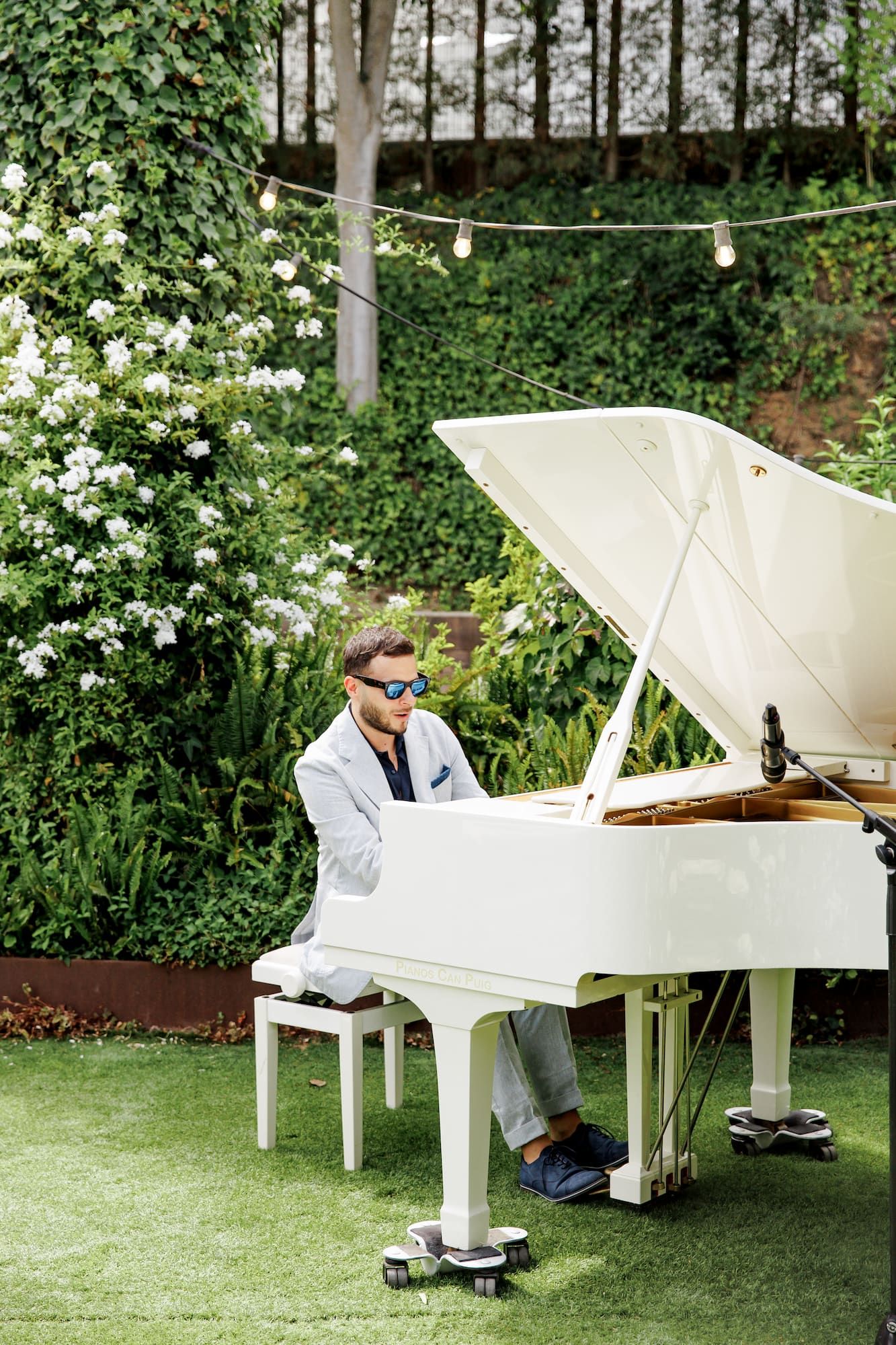 Man in white suit playing a white grand piano outdoors in a lush garden setting with string lights.