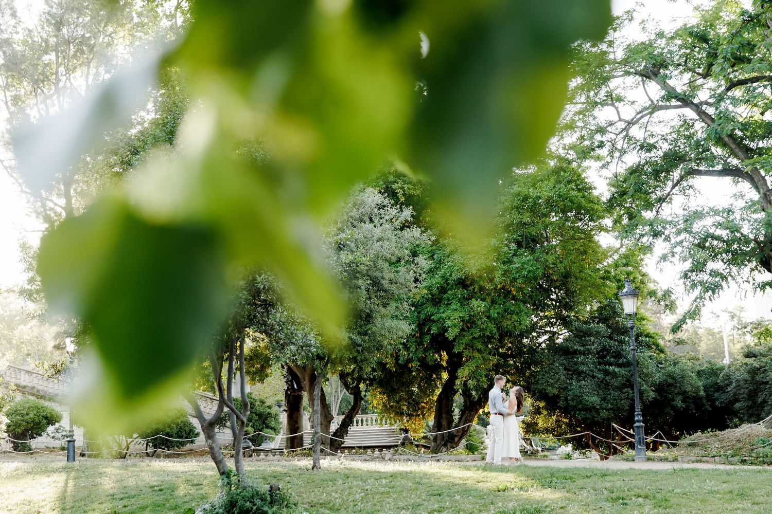 Couple stands in lush green garden surrounded by tall trees, captured through soft bokeh leaves in foreground.