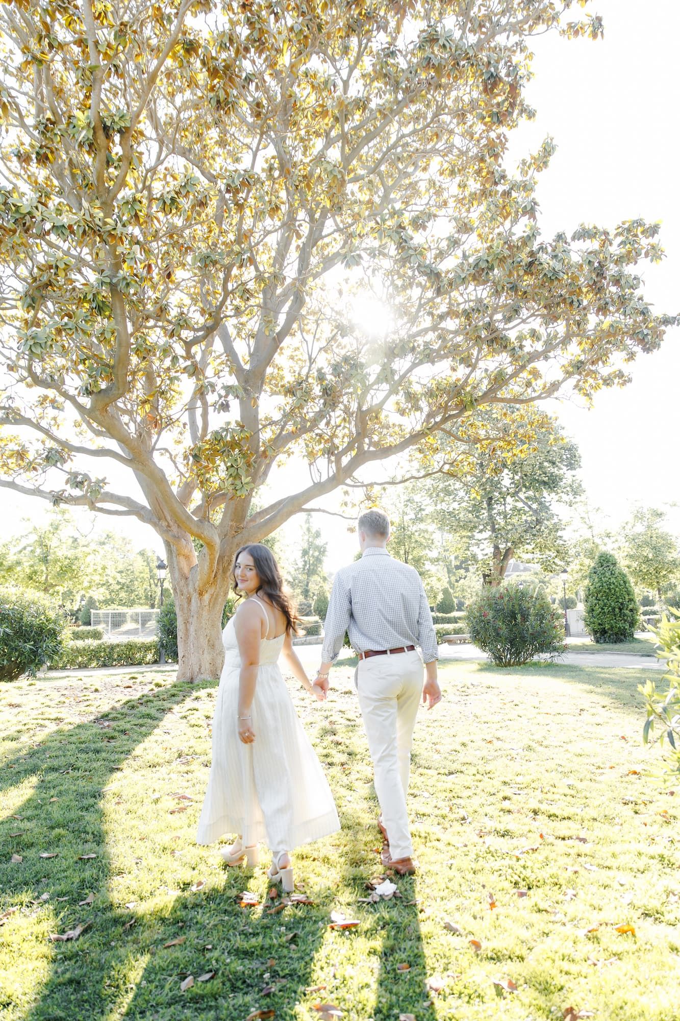 Couple holding hands walking in sunlit park, woman in white dress glancing back, man in light gray outfit near large tree.