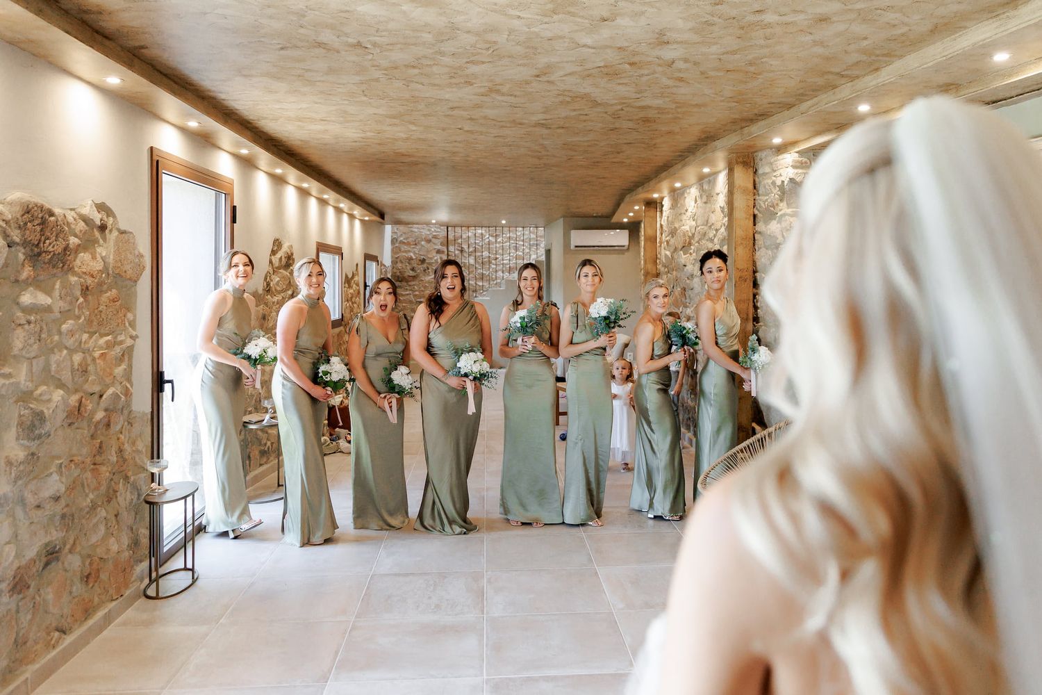 Bridesmaids in sage green gowns holding white bouquets smile at bride during wedding day first look.