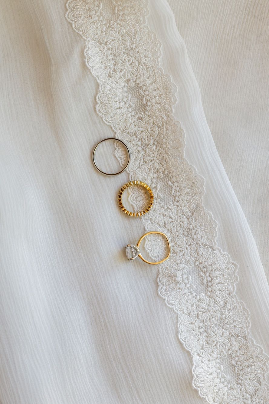 Three wedding rings, including a gold solitaire engagement ring, laid on white fabric beside delicate floral lace trim.