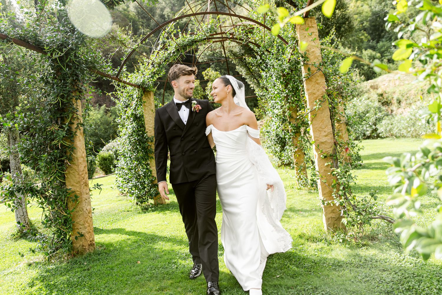 Bride and groom walk under lush green garden arch during a sunny day at Mas Huix, Barcelona