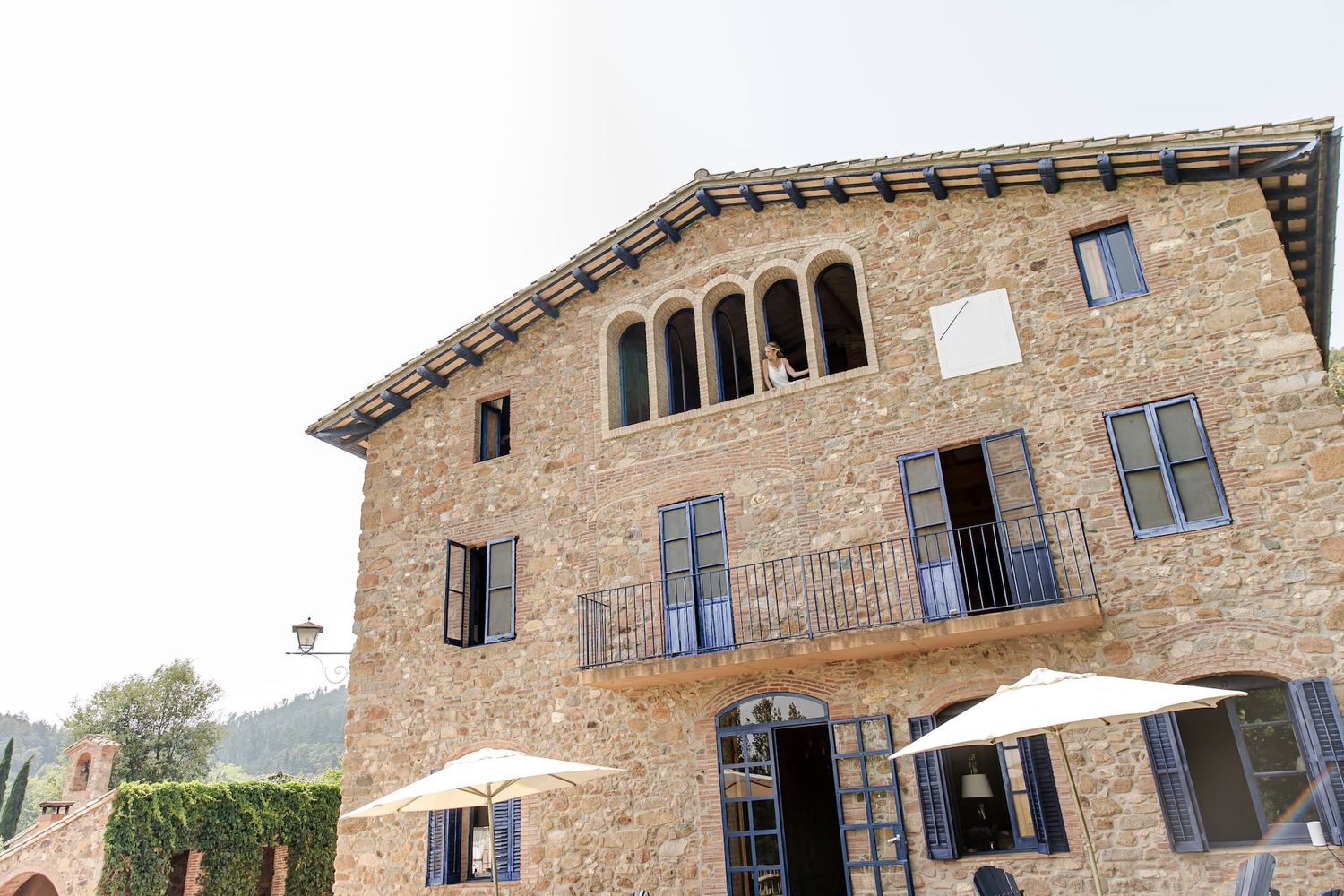 Rustic stone villa with arched windows, balcony, and white umbrellas surrounded by lush greenery.