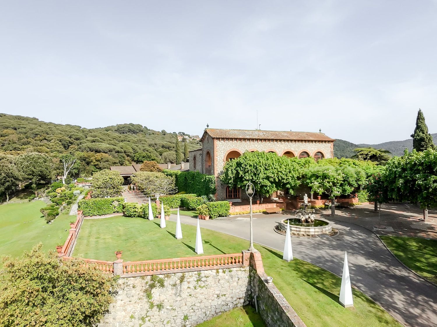 Aerial view of a wedding villa with manicured gardens, tall cypress trees, and a brick building surrounded by lush green hills.