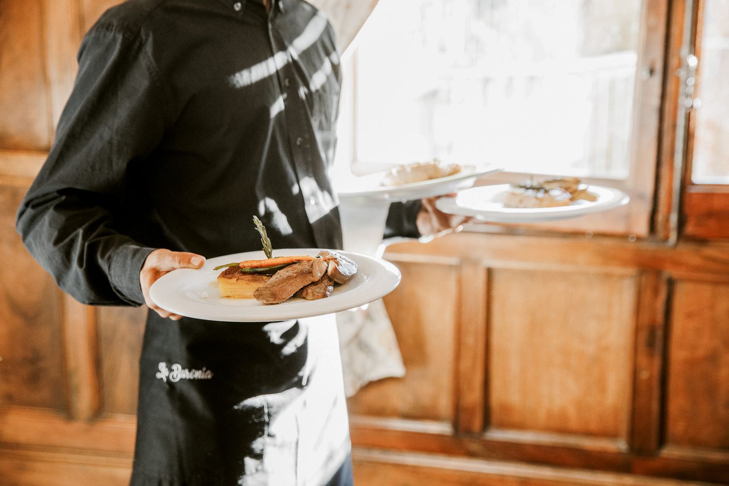 Waiter in black apron carrying multiple plates of food in the dining room of La Baronia