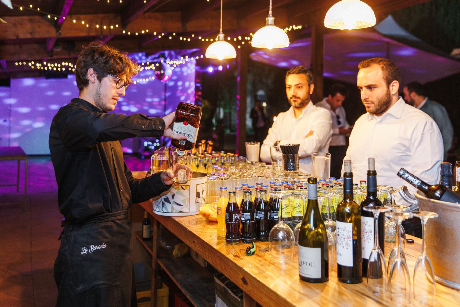 Bartender pouring drinks at a well-stocked bar with wine bottles, serving two guests at an elegant evening event.