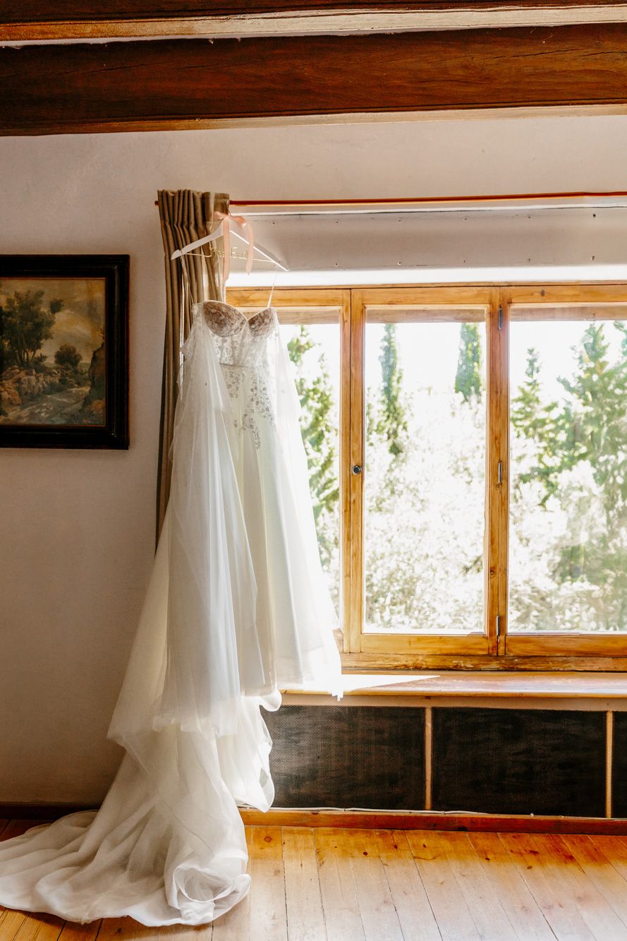 White wedding dress hanging on a wooden window frame in a rustic room with hardwood floors and natural light.