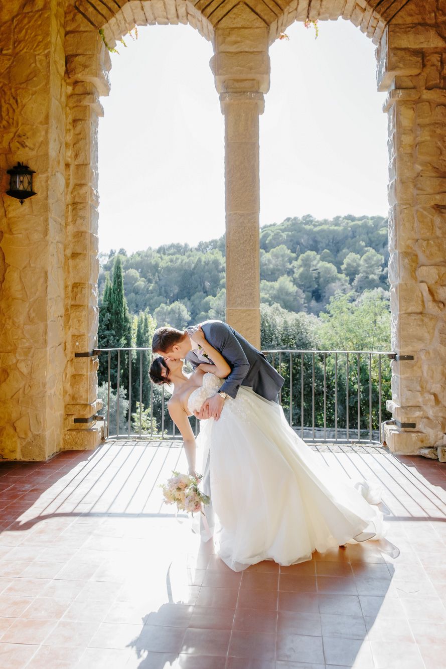 Bride and groom share a romantic dip kiss on a sunlit stone balcony of La Baronia wedding venue