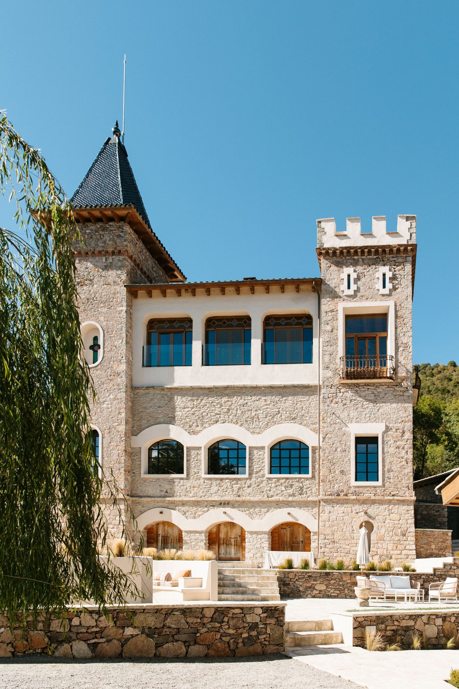Historic stone castle-style building with a crenellated tower, slate turret, arched blue windows, and a terrace under a clear blue sky.
