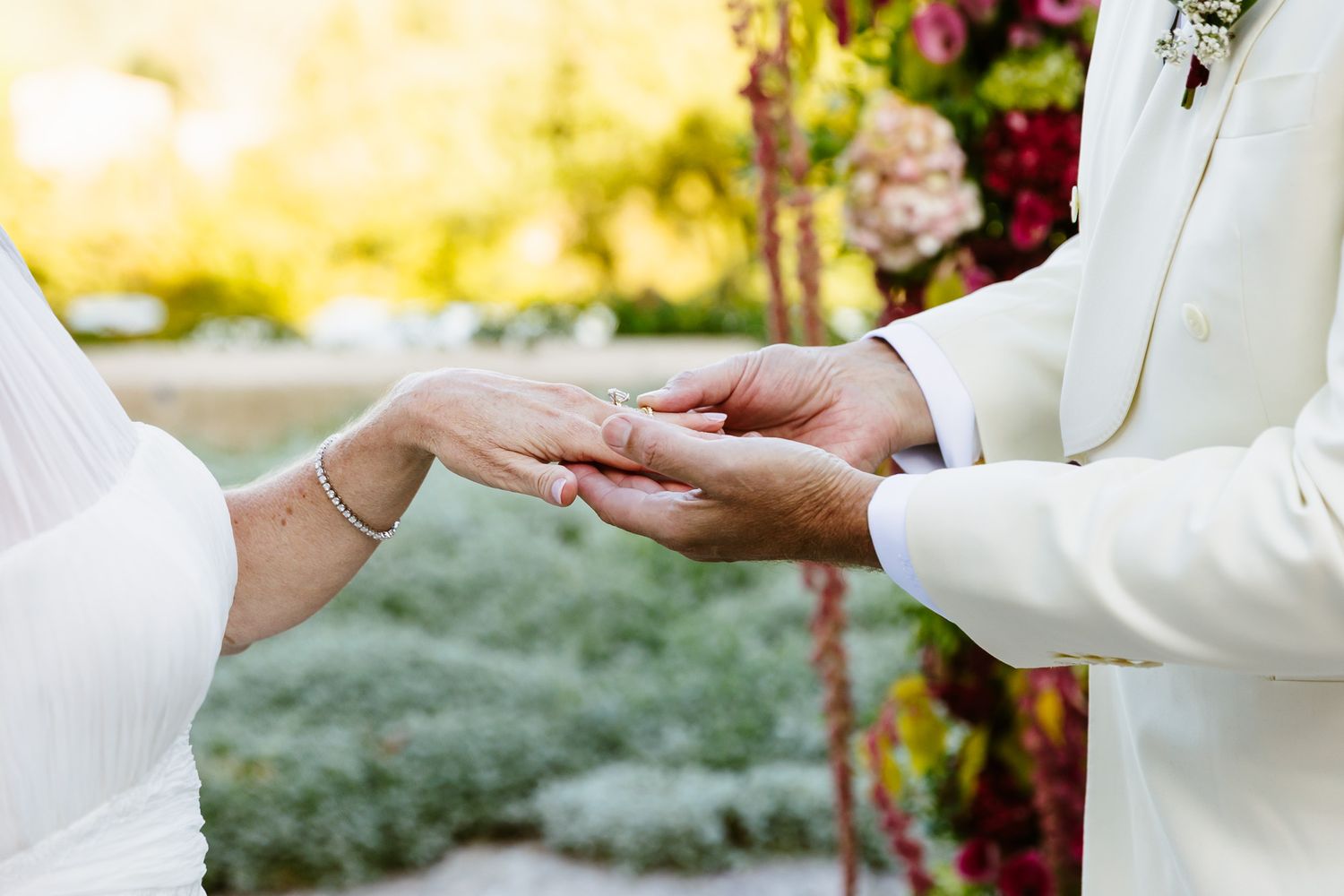 Bride in a white dress and groom in a cream suit hold hands during a wedding ring exchange in a garden with colorful flowers.