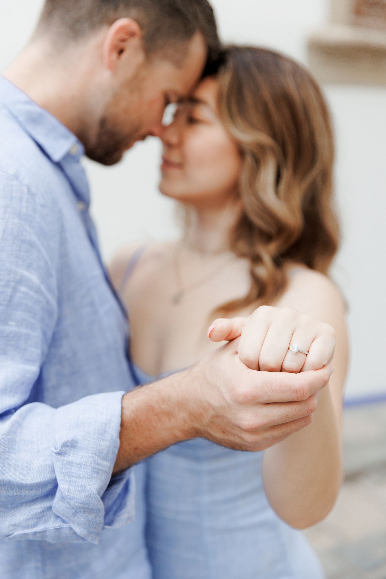 A romantic close-up of an engaged couple holding hands and touching foreheads, with a sparkling engagement ring visible.