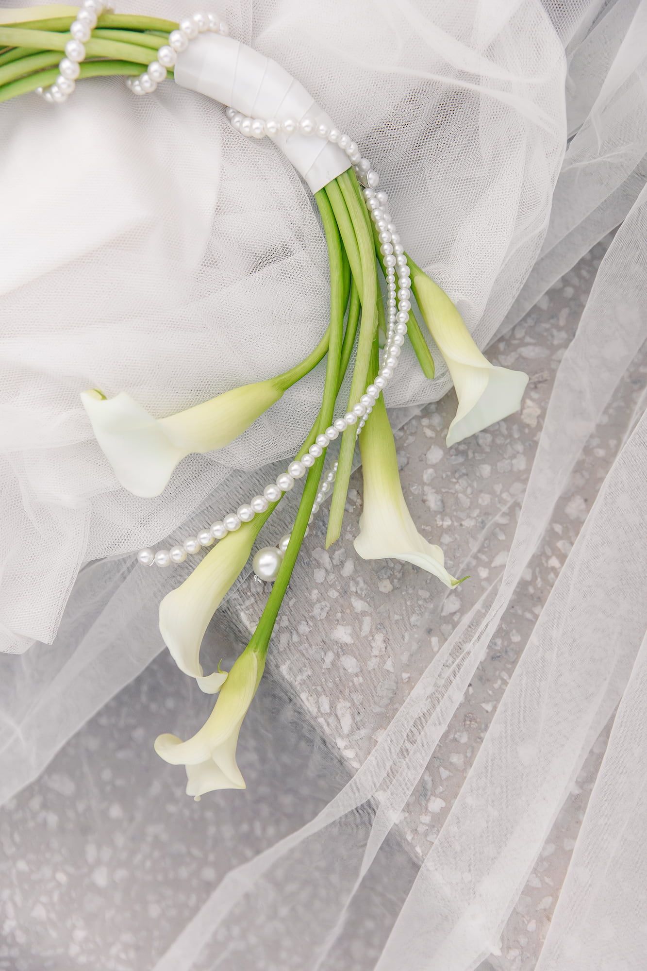 White calla lilies with green stems lay elegantly against flowing tulle fabric and delicate pearl strands.