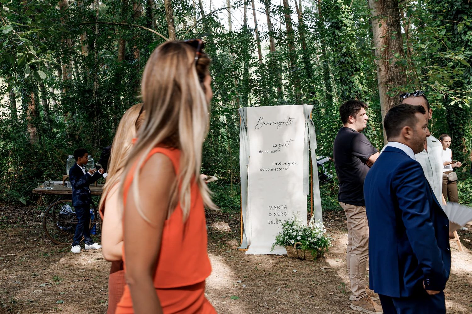 Outdoor wedding reception in a forest setting with guests near a white welcome sign surrounded by greenery.