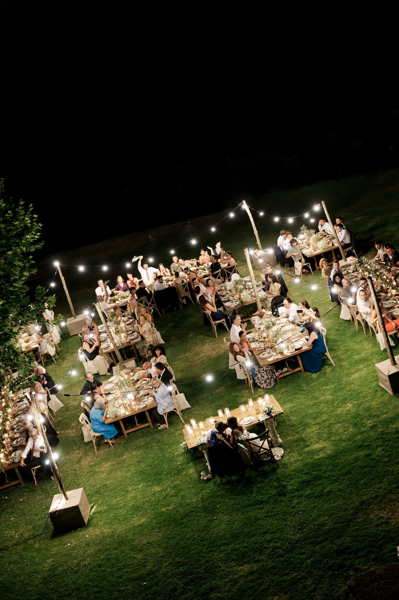 Aerial view of an elegant outdoor wedding reception at night with string lights illuminating guests dining on a green lawn.