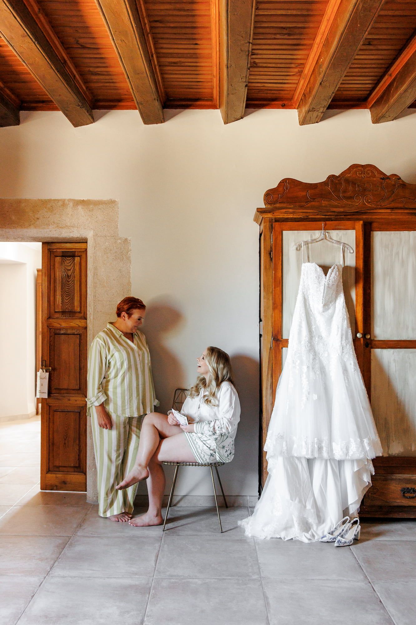 A white wedding dress hangs on an antique wooden wardrobe in a rustic room with exposed wooden beams.