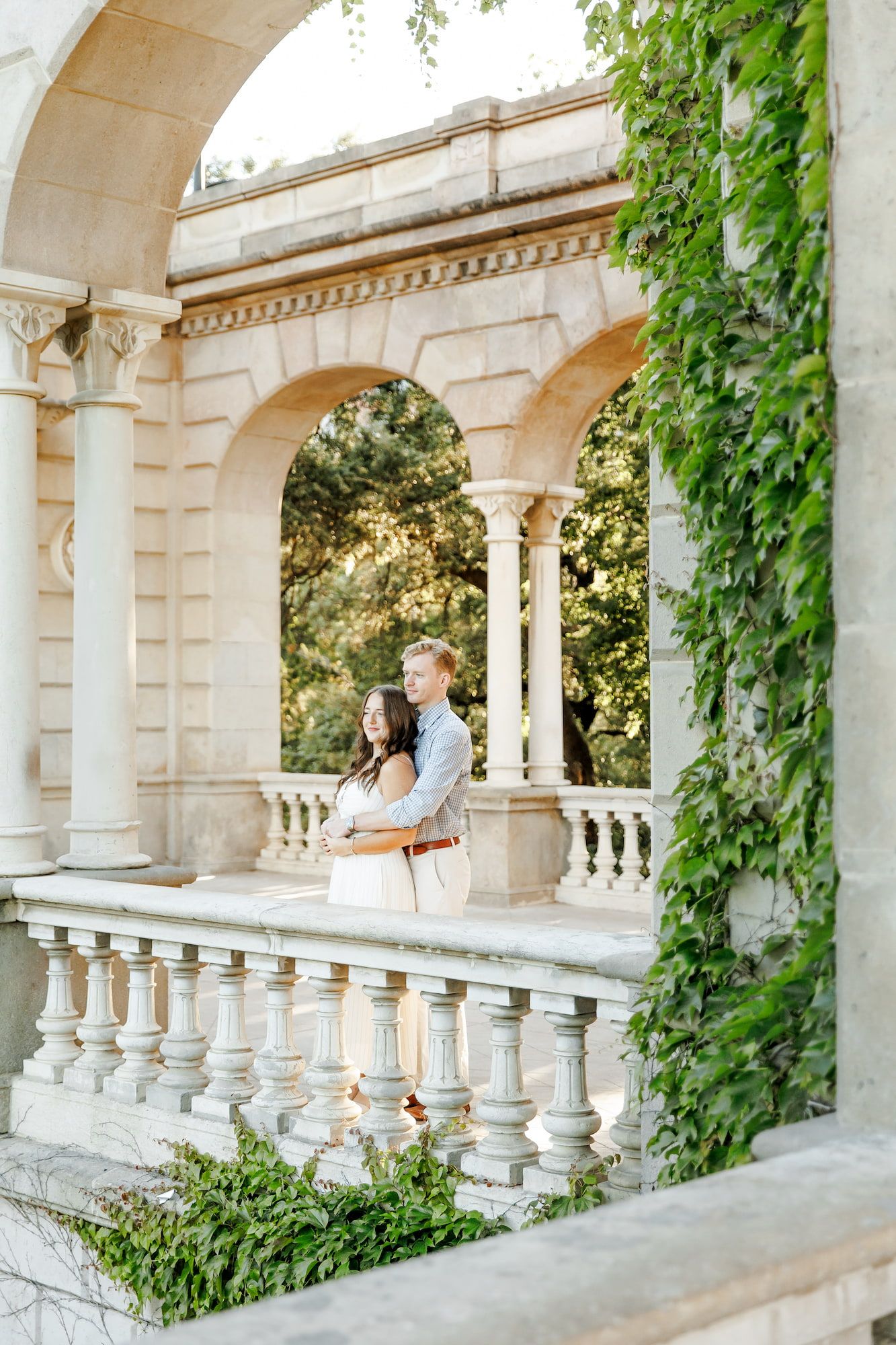 Couple embracing under classical stone arches with ivy, romantic engagement photo at elegant historic building.