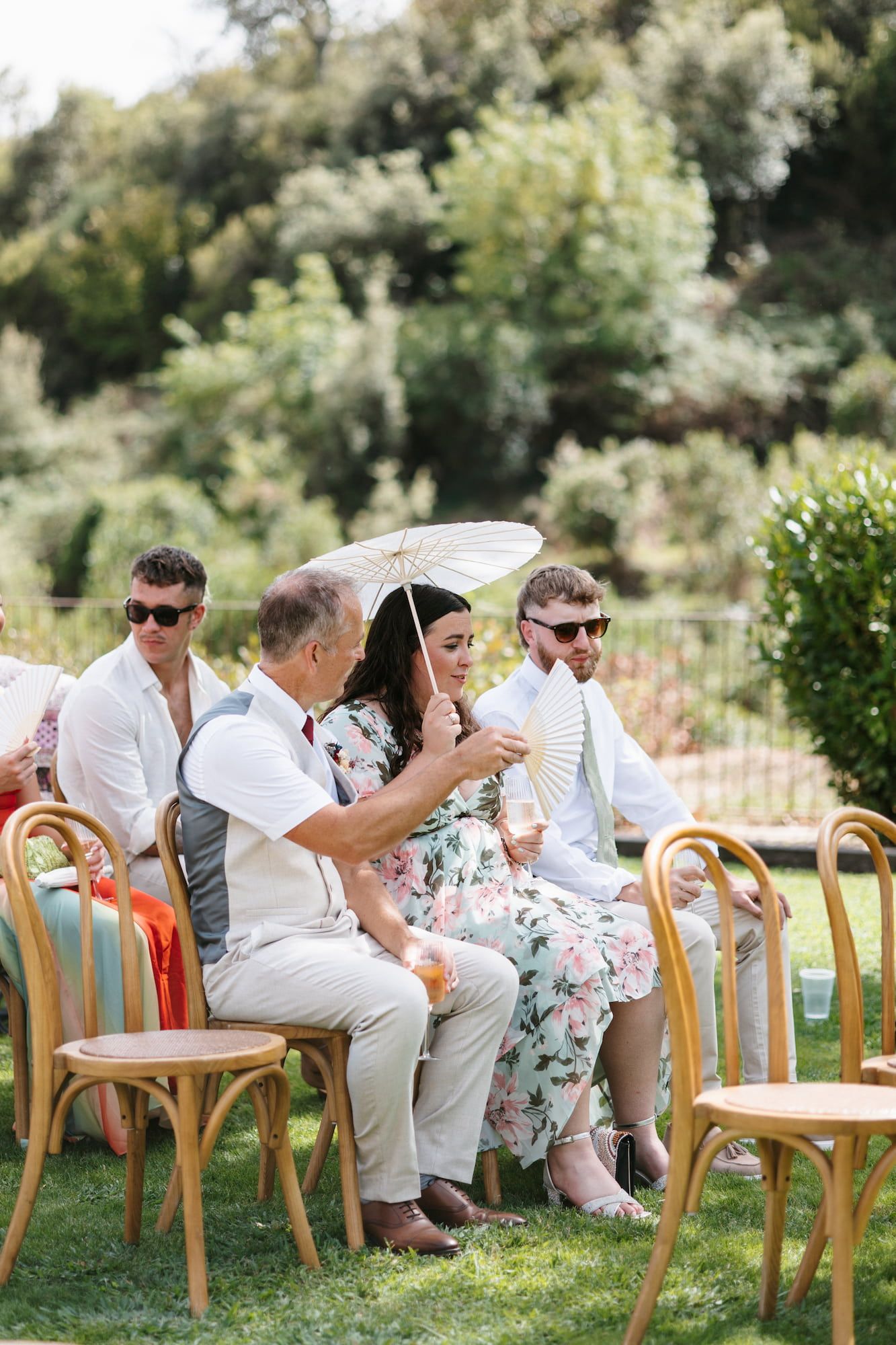 Wedding guests seated outdoors on wooden chairs, woman in floral dress holding parasol on sunny garden ceremony day.