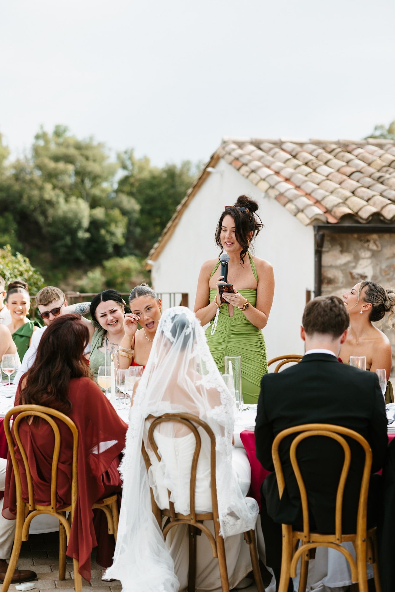 Bridesmaid in green dress gives speech at outdoor wedding reception as bride and guests listen attentively.