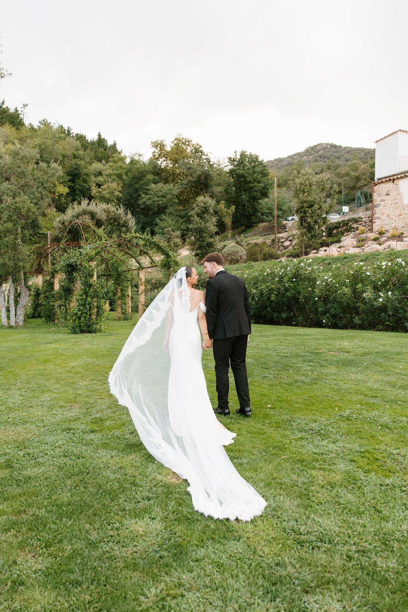 Bride and groom share a kiss outdoors on lush green lawn, her cathedral veil flowing in the breeze with garden backdrop.